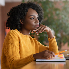 Thoughtful afro young woman looking for new job online, using laptop at cafe, taking notes, side view, copy space