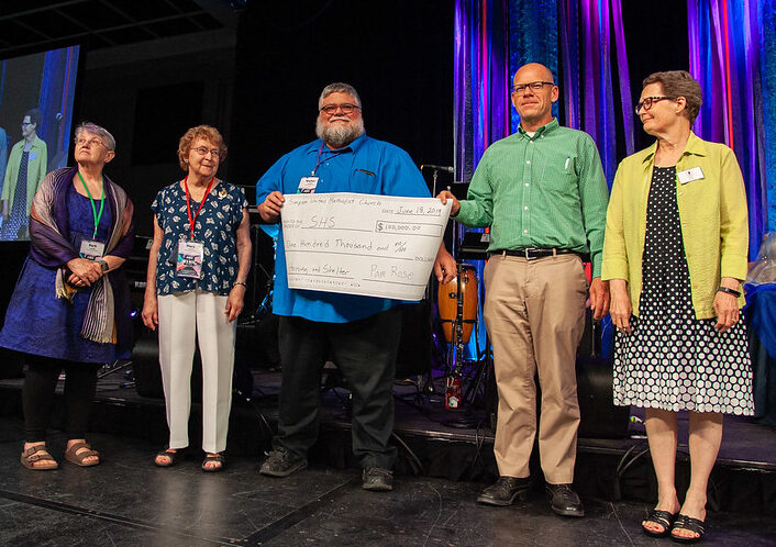 Five people stand on a stage, two of them holding a large check.
