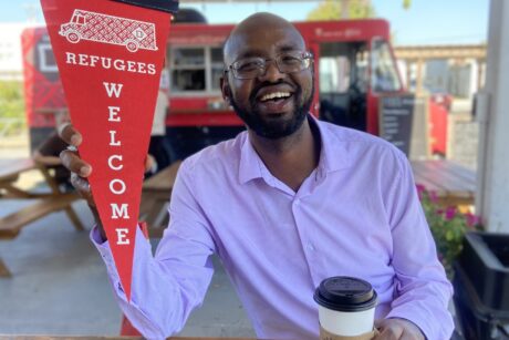 A man smiles at the camera while sitting at a picnic bench holding a coffee cup in one hand and a pendant that reads "Refugees Welcome" in the other. Behind him is a red Refuge Coffee Co. truck.