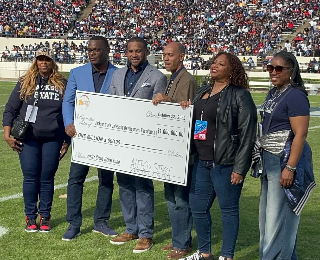 Representatives form Jackson State University and Alfred Street Baptist stand on the turf during a football game holding a large check.