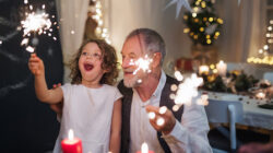 Senior grandfather with small granddaughter indoors at Christmas, sitting at table with sparklers.