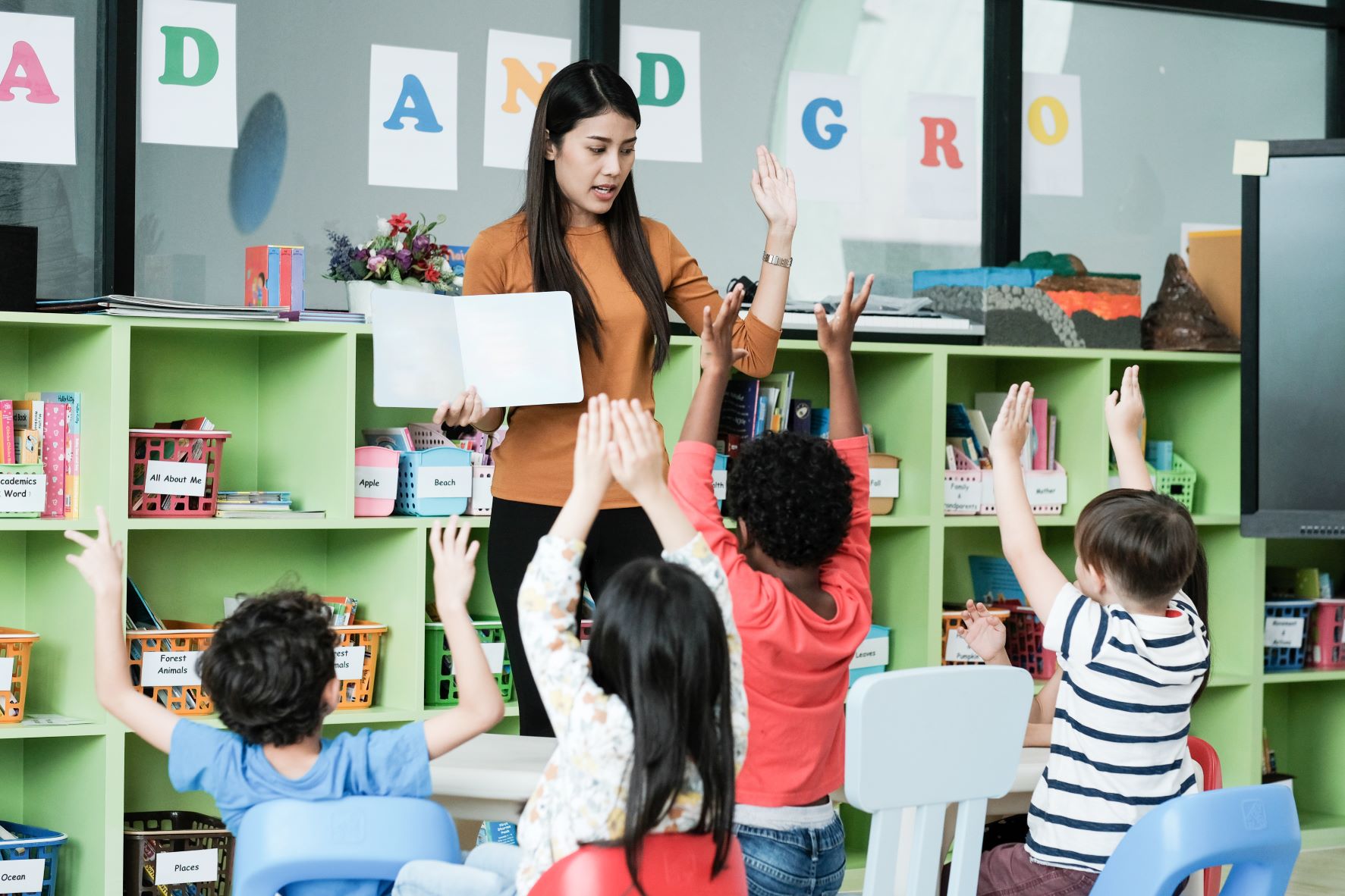 Photo of a teacher facing kindergarden students in a classroom. The students have their hands up as the teacher readers them a book.