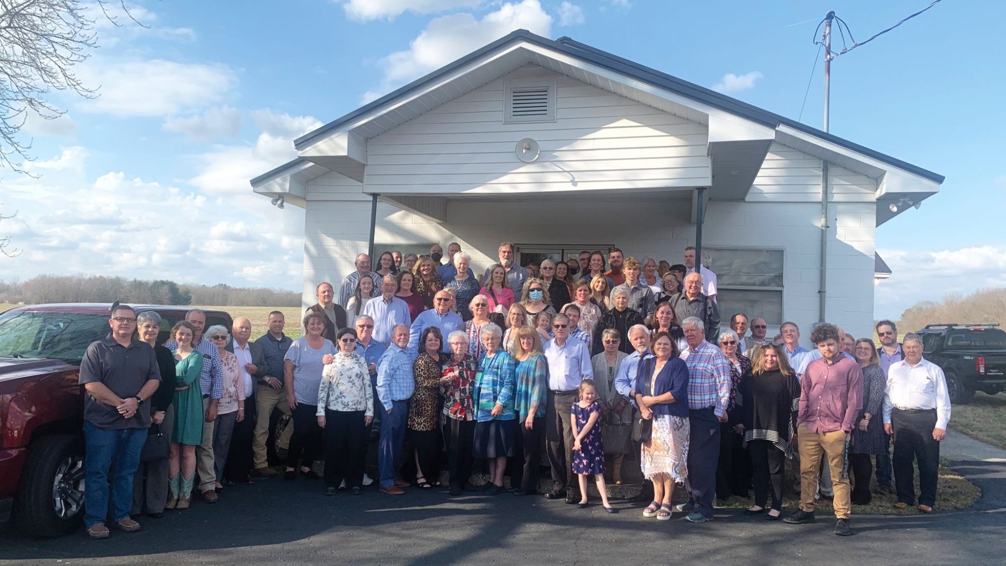 Photo of about 50 people dressed for a church service in front a small church surrounded by open fields.
