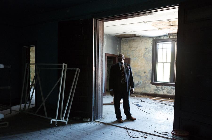 A pastor in a suit stands in a dilapidated building.