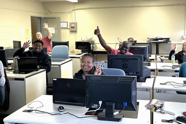Students sit in a classroom behind computers, smiling at the camera and giving thumbs up.