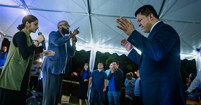 An African-American pastor prays into a microphone next to a Latina interpreter holding a microphone while a Latino preacher holds his hands up in prayer.