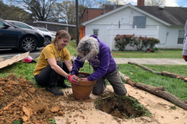 Two women kneel on the ground while putting a plant in a planter next to a hole in the ground.