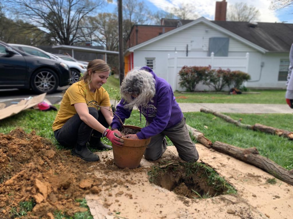Two women kneel on the ground while putting a plant in a planter next to a hole in the ground.