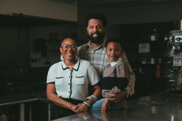 A man, wife, and child stand smiling in a commercial kitchen.