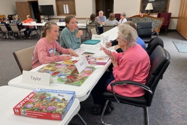 Youth and elderly folks sit across from one another working on a puzzle and talking.