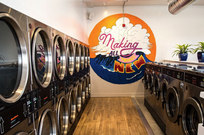 Photo of laundry machines with plants on top and a wall mural that reads: Making All Things New