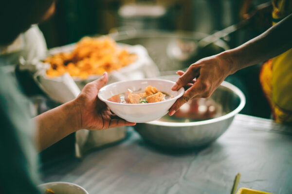 Person hands a bowl of soup to another person, with large bowls of food in the background