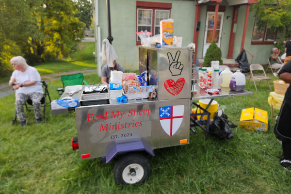Photo of a hot dog cart that says "Feed My Sheep Ministries" and has the Episcopal Church logo on it. Various food items, plates, and utensils sit on top of it. It's parked on the grass and people are standing and sitting around the cart.