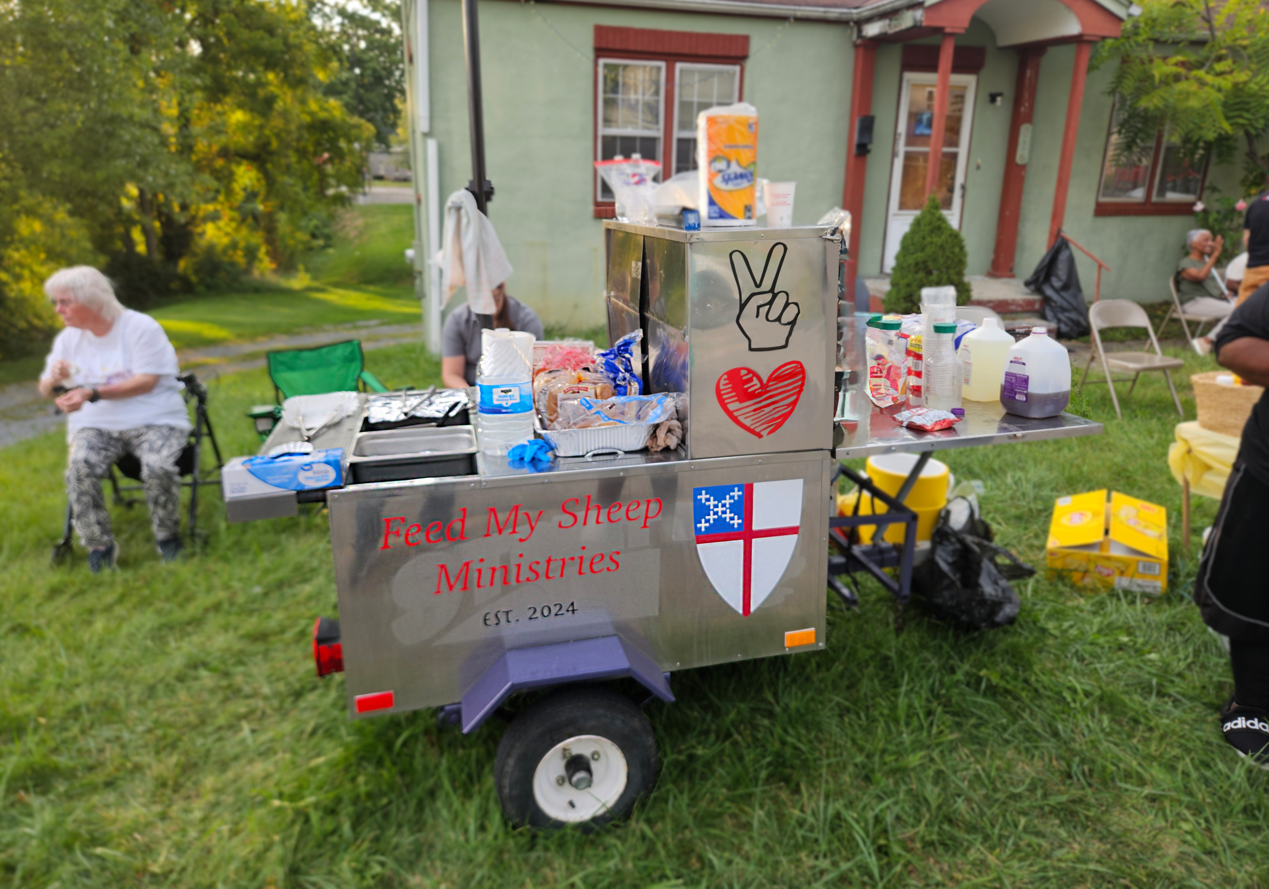 Photo of a hot dog cart that says "Feed My Sheep Ministries" and has the Episcopal Church logo on it. Various food items, plates, and utensils sit on top of it. It's parked on the grass and people are standing and sitting around the cart.