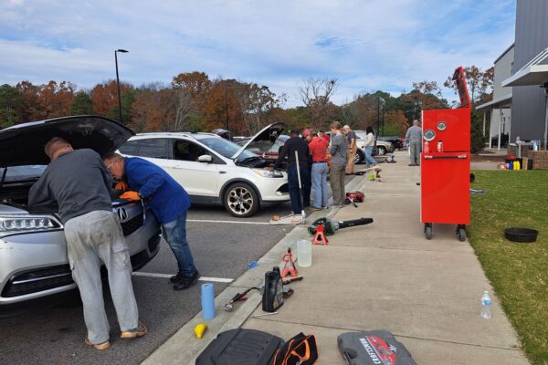Photo of several cars in a church parking lot with mechanics and owners looking at the engines as large toolboxes surround them.