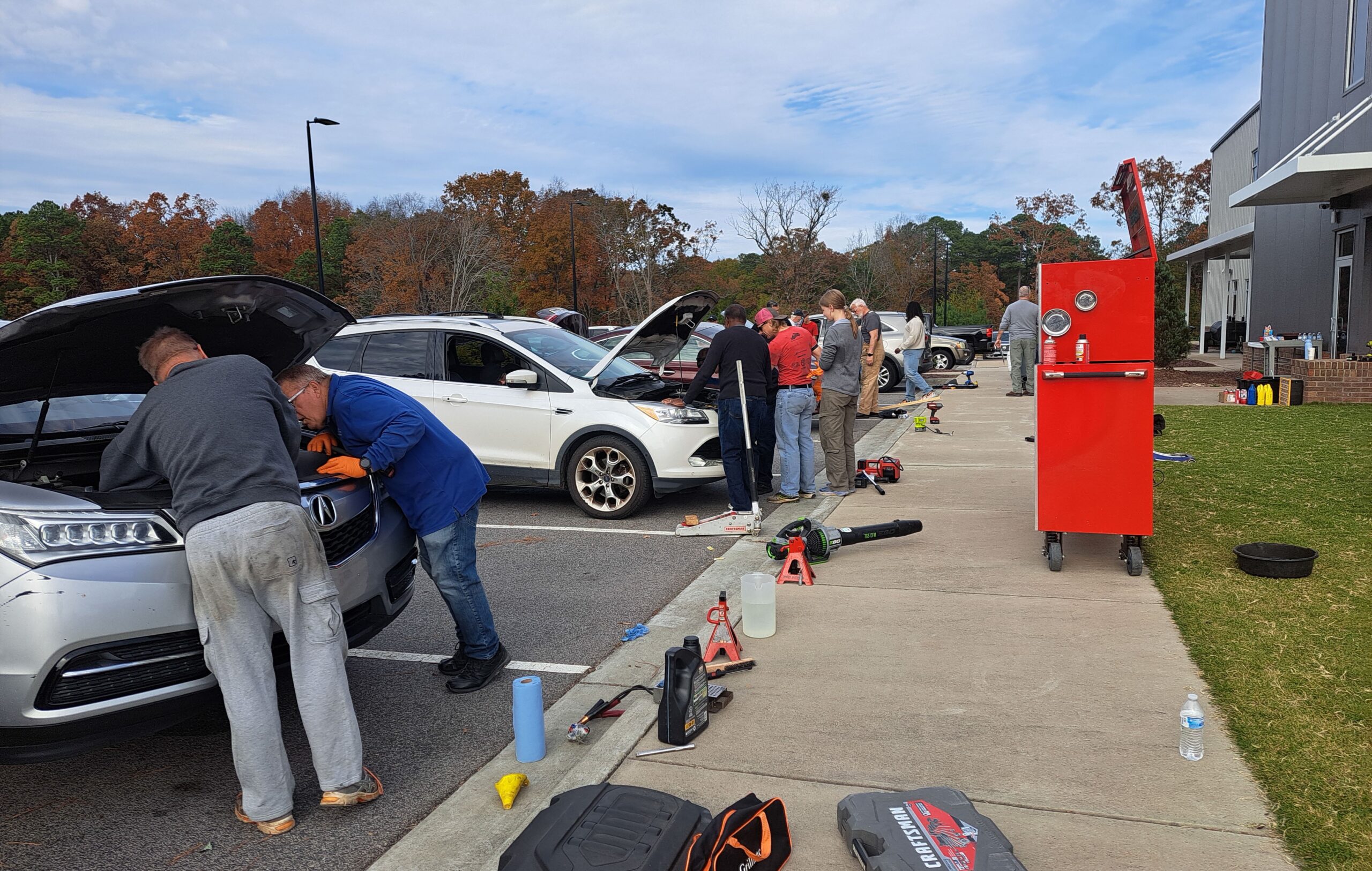 Photo of several cars in a church parking lot with mechanics and owners looking at the engines as large toolboxes surround them.