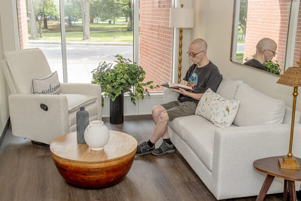 An elderly man sits on a sofa in a bright, modern living room, reading a book. Large windows overlook a tree-lined street.