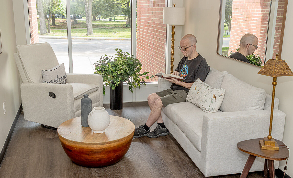 An elderly man sits on a sofa in a bright, modern living room, reading a book. Large windows overlook a tree-lined street.