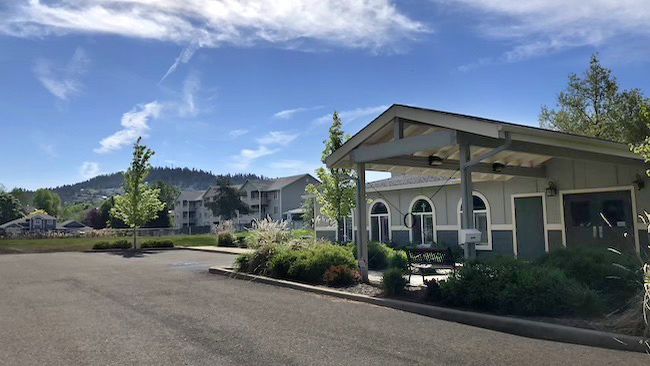 Exterior view of a small covered pavilion and office building with arched windows, in a residential neighborhood, with forested hills and blue sky in the background