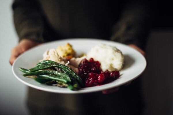 A stock photo of a person holding out a white plate filled with a Thanksgiving meal, including sliced turkey, stuffing, mashed potatoes, cranberry sauce, and green beans.