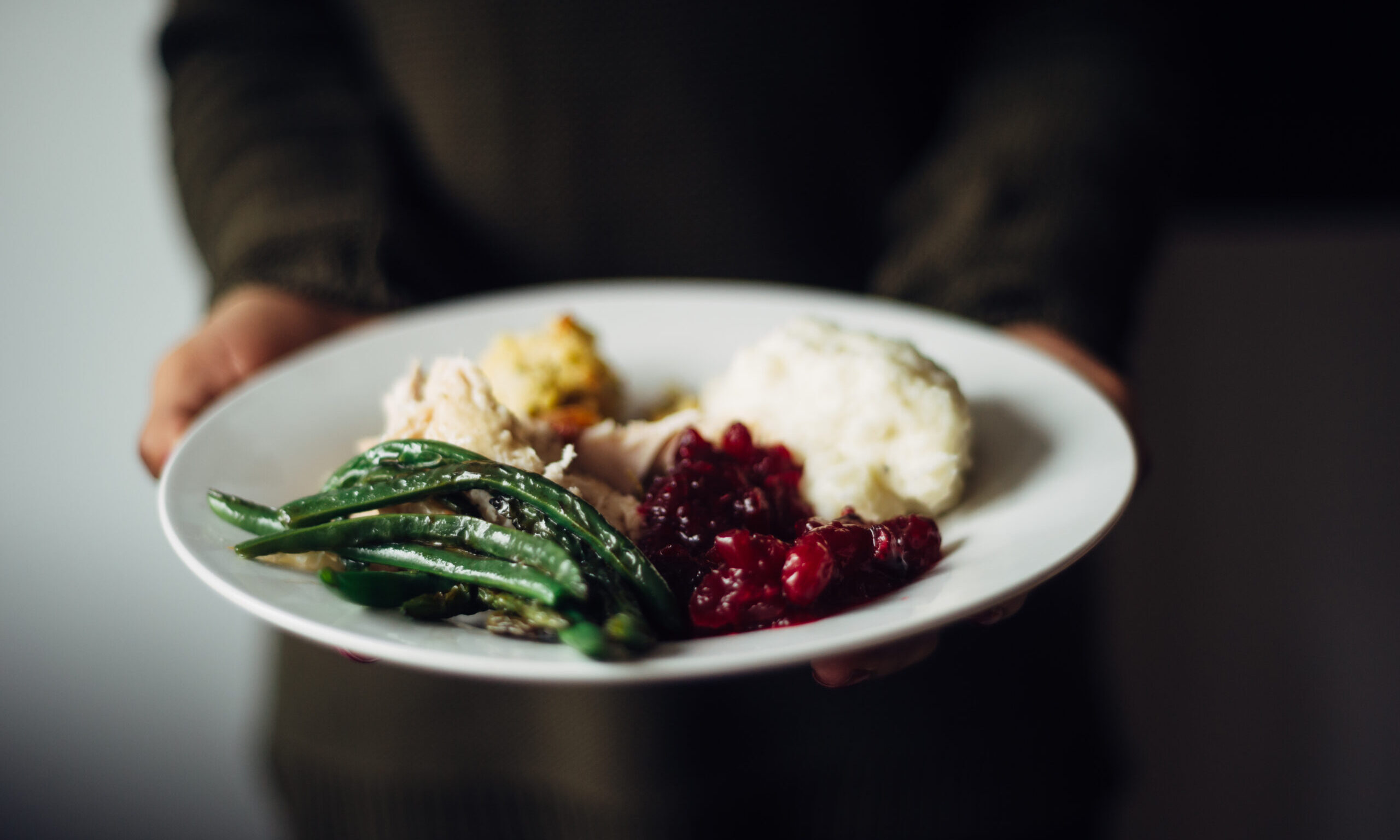 A stock photo of a person holding out a white plate filled with a Thanksgiving meal, including sliced turkey, stuffing, mashed potatoes, cranberry sauce, and green beans.