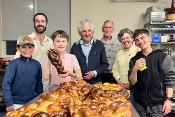A group of two men, two boys, a woman, and an older couple stand behind a table with dozens of freshly baked, glossy braided challah loaves. One boy holds up a single braided loaf for the camera. Everyone is smiling in what appears to be an industrial kitchen.