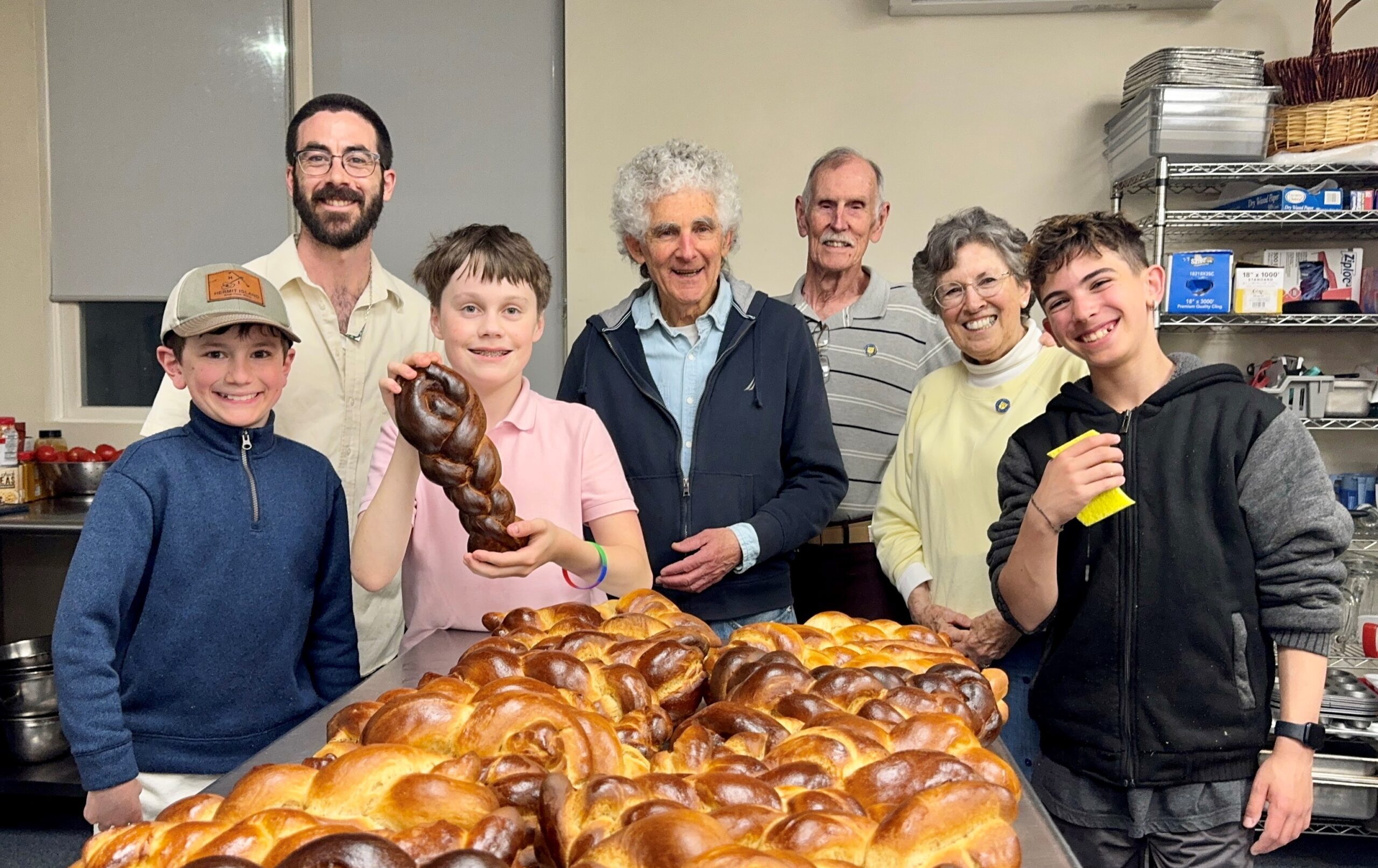 A group of two men, two boys, a woman, and an older couple stand behind a table with dozens of freshly baked, glossy braided challah loaves. One boy holds up a single braided loaf for the camera. Everyone is smiling in what appears to be an industrial kitchen.